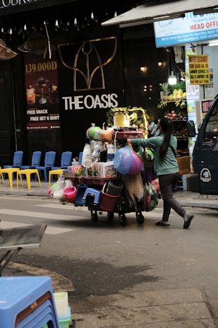 A street vendor is pushing a cart loaded with various household goods. The cart is stocked with colorful plastic baskets, brooms, and other miscellaneous items. The background features a store with a sign reading 'THE OASIS' and vibrant yellow and blue chairs are lined up outside.
