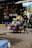 A street vendor is pushing a cart loaded with various household goods. The cart is stocked with colorful plastic baskets, brooms, and other miscellaneous items. The background features a store with a sign reading 'THE OASIS' and vibrant yellow and blue chairs are lined up outside.