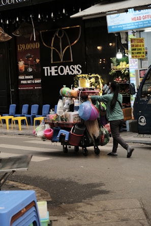A street vendor is pushing a cart loaded with various household goods. The cart is stocked with colorful plastic baskets, brooms, and other miscellaneous items. The background features a store with a sign reading 'THE OASIS' and vibrant yellow and blue chairs are lined up outside.