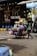 A street vendor is pushing a cart loaded with various household goods. The cart is stocked with colorful plastic baskets, brooms, and other miscellaneous items. The background features a store with a sign reading 'THE OASIS' and vibrant yellow and blue chairs are lined up outside.