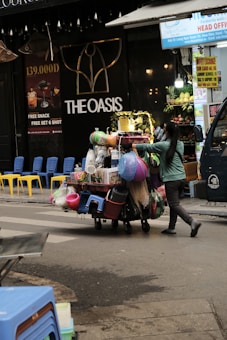 A street vendor is pushing a cart loaded with various household goods. The cart is stocked with colorful plastic baskets, brooms, and other miscellaneous items. The background features a store with a sign reading 'THE OASIS' and vibrant yellow and blue chairs are lined up outside.