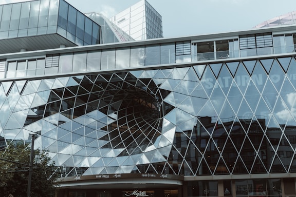 A modern building facade featuring a complex, geometrically-patterned glass structure. The glass panels create a reflective surface, with a large, eye-catching, funnel-like void in the center. The design includes numerous triangular and diamond shapes, mirroring the sky and surrounding urban environment. Signs for various businesses, including 'Saturn' and 'Foodtopia', are visible through the glass.