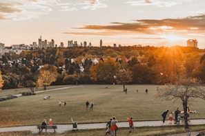 A scenic park landscape during sunset with people enjoying recreational activities. In the background, a city skyline is visible with tall buildings, while the foreground features a spacious grassy field surrounded by autumn foliage. Several groups of people are seen walking, sitting, and playing in the park, adding a sense of liveliness to the serene setting.