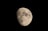 A detailed view of the moon is captured with its surface illuminated, showing various craters and rugged terrain against a dark black sky.
