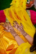 Intricately designed henna patterns are applied on both hands, which are held out on a vibrant yellow and orange fabric. There are also marigold flowers and a decorative pot in the background.