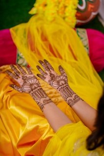 Intricately designed henna patterns are applied on both hands, which are held out on a vibrant yellow and orange fabric. There are also marigold flowers and a decorative pot in the background.