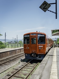 An orange diesel train is positioned at a small rural train station under a clear blue sky. The train tracks are visible, and a sign with the number '2' hangs above the platform. In the background, there are green grass and trees, and part of the station platform is visible.