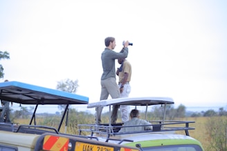 Two individuals are standing on the rooftop of a safari vehicle. One appears to be using a camera or binoculars, suggesting they are observing wildlife. The scene is set in an open grassland with sparse trees visible in the background, indicative of a savanna environment.