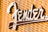 A close-up view of a metallic logo attached to a textured fabric background. The logo features a stylized font in a shiny metallic finish.