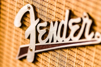 A close-up view of a metallic logo attached to a textured fabric background. The logo features a stylized font in a shiny metallic finish.