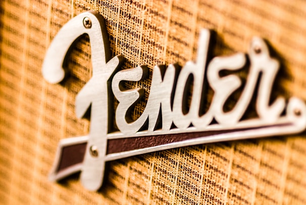 A close-up view of a metallic logo attached to a textured fabric background. The logo features a stylized font in a shiny metallic finish.