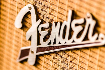 A close-up view of a metallic logo attached to a textured fabric background. The logo features a stylized font in a shiny metallic finish.