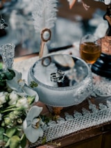 Delicate wedding rings are placed on an ornate glass bowl surrounded by elegant lace and greenery. A crystal goblet filled with amber liquid and white flowers add to the sophisticated setting.