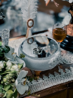 Delicate wedding rings are placed on an ornate glass bowl surrounded by elegant lace and greenery. A crystal goblet filled with amber liquid and white flowers add to the sophisticated setting.