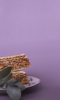Crispy layered wafer slices arranged on a decorative plate with floral patterns. A sprig of eucalyptus leaves rests partially covering the bottom of the stack, against a soft purple backdrop.