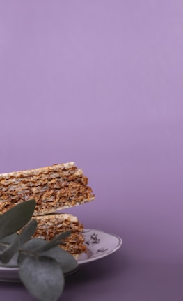 Crispy layered wafer slices arranged on a decorative plate with floral patterns. A sprig of eucalyptus leaves rests partially covering the bottom of the stack, against a soft purple backdrop.