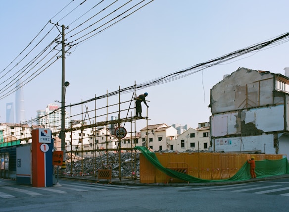 A construction area with scaffolding and debris, surrounded by partially demolished buildings. A person is visible working on the scaffolding, while another individual dressed in an orange outfit is near a temporary barrier. Urban buildings and a distinctive modern skyscraper can be seen in the background. Power lines and poles are also visible.