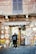 A person stands in front of a rustic shop with a brick facade, displaying a variety of colorful ceramic plates and postcards. The shop sign reads 'Antica Siena'. The door is made of glass, and the person is wearing a dark jacket and a blue backpack.