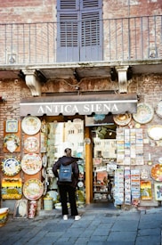 A person stands in front of a rustic shop with a brick facade, displaying a variety of colorful ceramic plates and postcards. The shop sign reads 'Antica Siena'. The door is made of glass, and the person is wearing a dark jacket and a blue backpack.
