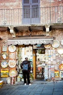 A person stands in front of a rustic shop with a brick facade, displaying a variety of colorful ceramic plates and postcards. The shop sign reads 'Antica Siena'. The door is made of glass, and the person is wearing a dark jacket and a blue backpack.