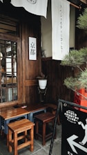A cozy corner featuring wooden furniture against a dark wooden wall, with small stools and a table. White fabric banners with Japanese characters hang overhead. A small potted plant adds a touch of greenery. A black sandwich board sign with 'Kyoto Country Club' and a silhouetted figure is visible in the foreground.