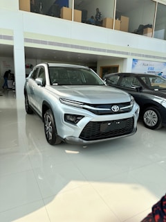A silver SUV is prominently displayed in a well-lit showroom with glossy tiled floors. The vehicle features sleek, modern design elements including a distinctive grille and sharp headlights. Other cars are visible alongside, and there are people present in an upstairs area visible through a large glass partition.