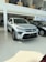 A silver SUV is prominently displayed in a well-lit showroom with glossy tiled floors. The vehicle features sleek, modern design elements including a distinctive grille and sharp headlights. Other cars are visible alongside, and there are people present in an upstairs area visible through a large glass partition.