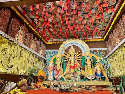 A richly decorated indoor setting featuring a vibrant Durga Puja pandal. The central focus is a large and ornate Durga idol with multiple arms, flanked by other deities. The background and ceiling are adorned with intricate patterns and red hanging decorations. Two people are seated on the left, possibly participating in a ritual.