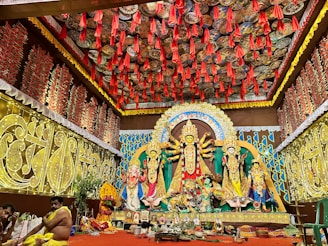 A richly decorated indoor setting featuring a vibrant Durga Puja pandal. The central focus is a large and ornate Durga idol with multiple arms, flanked by other deities. The background and ceiling are adorned with intricate patterns and red hanging decorations. Two people are seated on the left, possibly participating in a ritual.