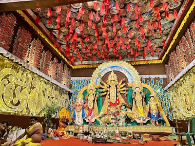 A richly decorated indoor setting featuring a vibrant Durga Puja pandal. The central focus is a large and ornate Durga idol with multiple arms, flanked by other deities. The background and ceiling are adorned with intricate patterns and red hanging decorations. Two people are seated on the left, possibly participating in a ritual.