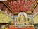 A richly decorated indoor setting featuring a vibrant Durga Puja pandal. The central focus is a large and ornate Durga idol with multiple arms, flanked by other deities. The background and ceiling are adorned with intricate patterns and red hanging decorations. Two people are seated on the left, possibly participating in a ritual.
