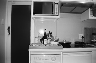 A kitchen counter with a dishwasher beneath it, displaying wine bottles and glasses, a wooden tray with prepared food, cutting boards, a microwave, and stovetop pans. The image is in black and white and features a modern kitchen setup with an open cupboard door.