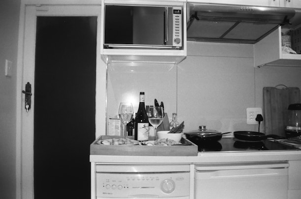A kitchen counter with a dishwasher beneath it, displaying wine bottles and glasses, a wooden tray with prepared food, cutting boards, a microwave, and stovetop pans. The image is in black and white and features a modern kitchen setup with an open cupboard door.