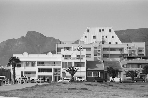 A black and white photograph featuring a combination of residential and commercial buildings in front of a mountain backdrop. The architecture includes modern and geometric designs, with visible palm trees lining the street. Vehicles are parked along the road, and a few people are walking through the area, suggesting an urban setting.