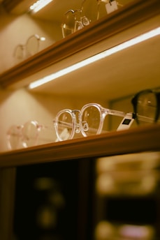 A collection of eyeglasses displayed on a well-lit shelf with a warm, ambient lighting effect. The image features multiple pairs of eyeglasses with transparent frames prominently in focus.