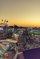 A vibrant carnival scene at sunset with numerous colorful stalls and rides. The area is bustling with people, and bright lights illuminate the carnival attractions. The sky is a gradient of warm colors, adding to the lively atmosphere.
