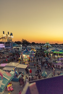 A vibrant carnival scene at sunset with numerous colorful stalls and rides. The area is bustling with people, and bright lights illuminate the carnival attractions. The sky is a gradient of warm colors, adding to the lively atmosphere.