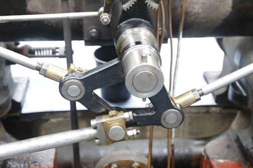 Close-up view of a metal mechanical component with interconnected rods and cylindrical gears, featuring metallic surfaces with various finishes, surrounded by industrial machinery parts.