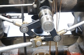 Close-up view of a metal mechanical component with interconnected rods and cylindrical gears, featuring metallic surfaces with various finishes, surrounded by industrial machinery parts.