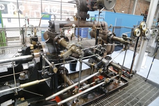 A large, intricate industrial steam engine is situated inside what appears to be an industrial setting. The engine is composed of numerous metallic rods, pistons, and gears, featuring a variety of brass and steel components. Gauges and pressure meters are attached, indicating monitoring measures. The background includes metal fencing and partial views of brick walls and windows.