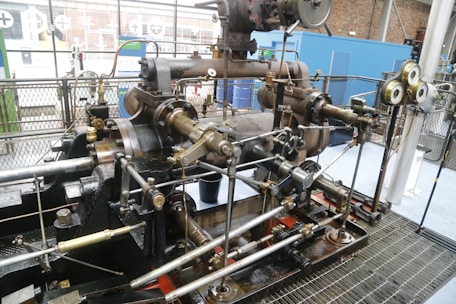 A large, intricate industrial steam engine is situated inside what appears to be an industrial setting. The engine is composed of numerous metallic rods, pistons, and gears, featuring a variety of brass and steel components. Gauges and pressure meters are attached, indicating monitoring measures. The background includes metal fencing and partial views of brick walls and windows.
