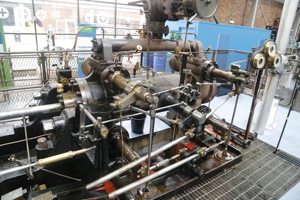 A large, intricate industrial steam engine is situated inside what appears to be an industrial setting. The engine is composed of numerous metallic rods, pistons, and gears, featuring a variety of brass and steel components. Gauges and pressure meters are attached, indicating monitoring measures. The background includes metal fencing and partial views of brick walls and windows.