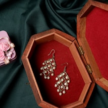A pair of pearl-studded earrings are elegantly displayed inside an open octagonal wooden box with a red velvet interior. The earrings have a chandelier design with an intricate arrangement of pearls. The background is a dark green fabric, and a small pink box with ribbon and rose decorations is visible on the left side.