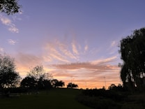 A picturesque sunset with vivid clouds stretches across the sky, casting a warm glow over a park landscape. Silhouettes of trees and lamp posts dot the horizon, adding depth to the scene. The sky transitions from deep blue at the top to soft oranges and pinks near the horizon, creating a tranquil and serene atmosphere.