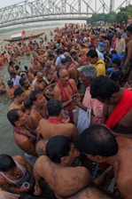 A large group of people gathered at a riverbank, participating in a religious or cultural ceremony. Many are partially submerged in water, wearing traditional attire. Diverse expressions grace their faces, from contemplative to joyous. The backdrop features a prominent steel bridge and lush greenery, suggesting an urban yet natural environment.