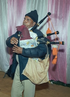 A person dressed in a dark jacket and hat is playing a bagpipe. The bagpipe appears to be made from various materials, and the musician is standing in front of a backdrop with pink and white fabric.