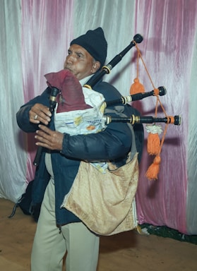 A person dressed in a dark jacket and hat is playing a bagpipe. The bagpipe appears to be made from various materials, and the musician is standing in front of a backdrop with pink and white fabric.