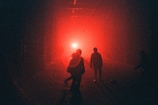 Silhouettes of people walk towards a bright red light in a dimly lit area, creating an atmosphere of mystery and intrigue.