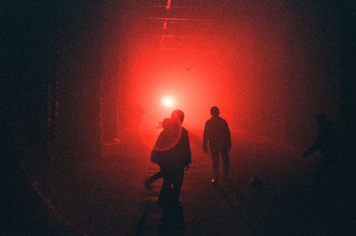 Silhouettes of people walk towards a bright red light in a dimly lit area, creating an atmosphere of mystery and intrigue.