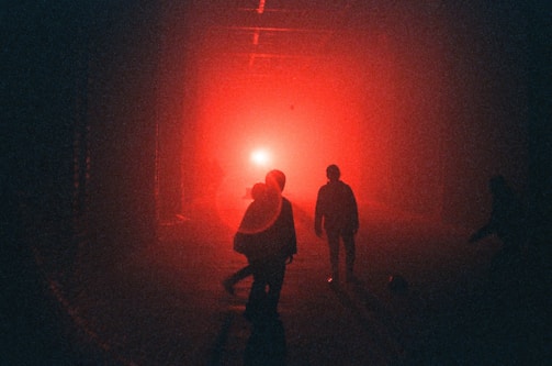 Silhouettes of people walk towards a bright red light in a dimly lit area, creating an atmosphere of mystery and intrigue.